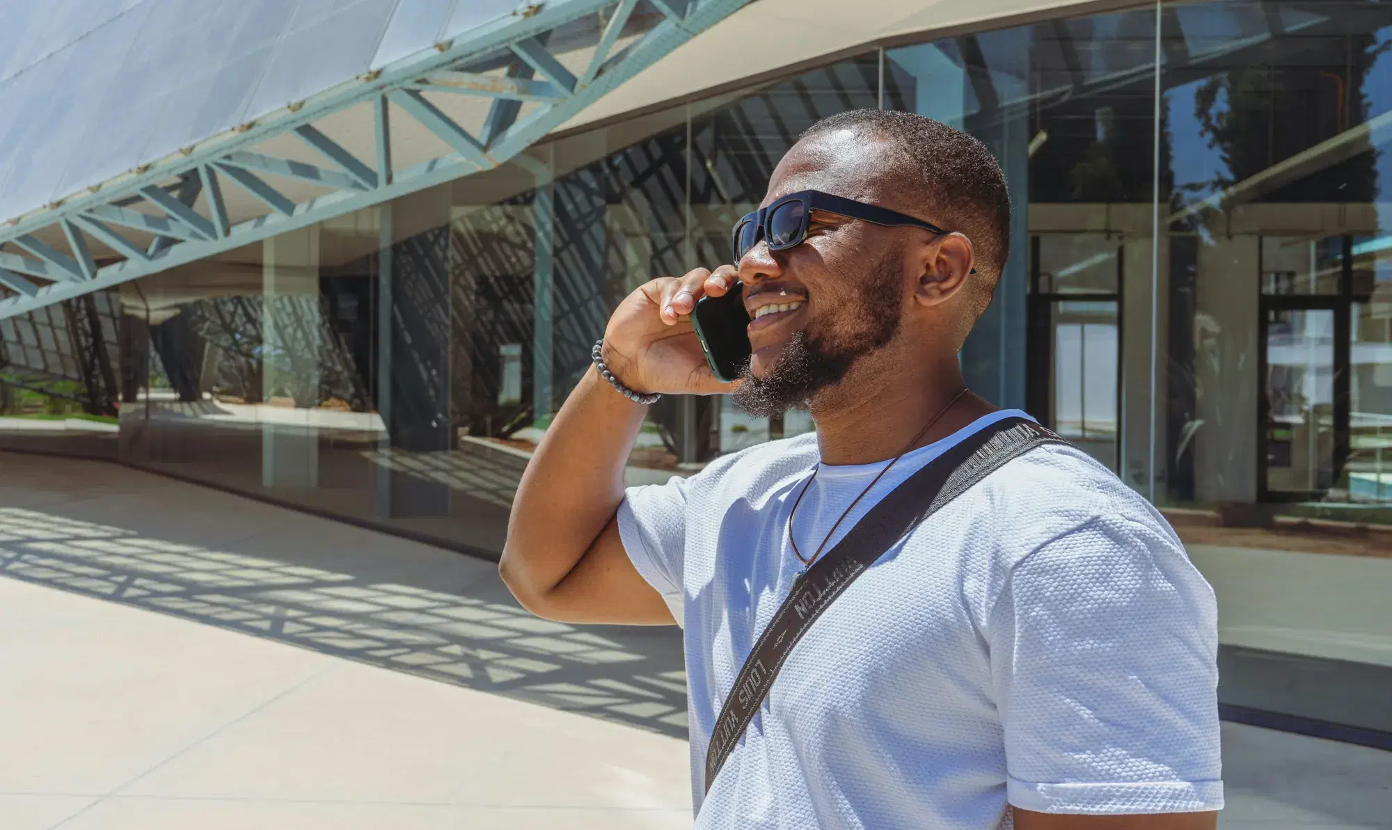 Homme souriant en lunettes de soleil téléphonant devant un bâtiment moderne vitré